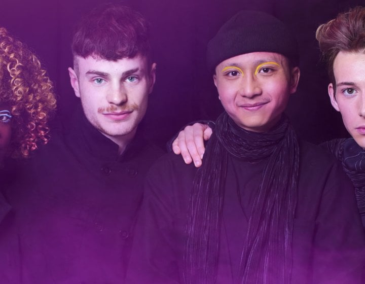 Photo of four student with their arms around each other's shoulders. Background is black with a pink haze.