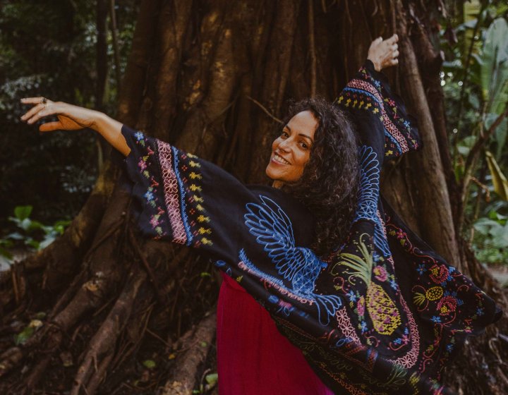 A performer in the woods in front od a tree with the arms in there air looking back over their shoulder towards the camera