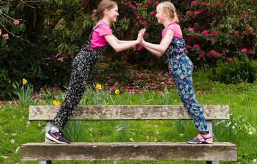 Two young dancers leaning towards each other on a bench in front of bushes and grass