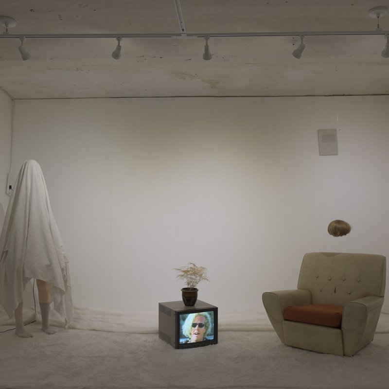 Room installation art, figure covered by white sheet, television on floor with woman's face, armchair, fan, wig suspended in the air.