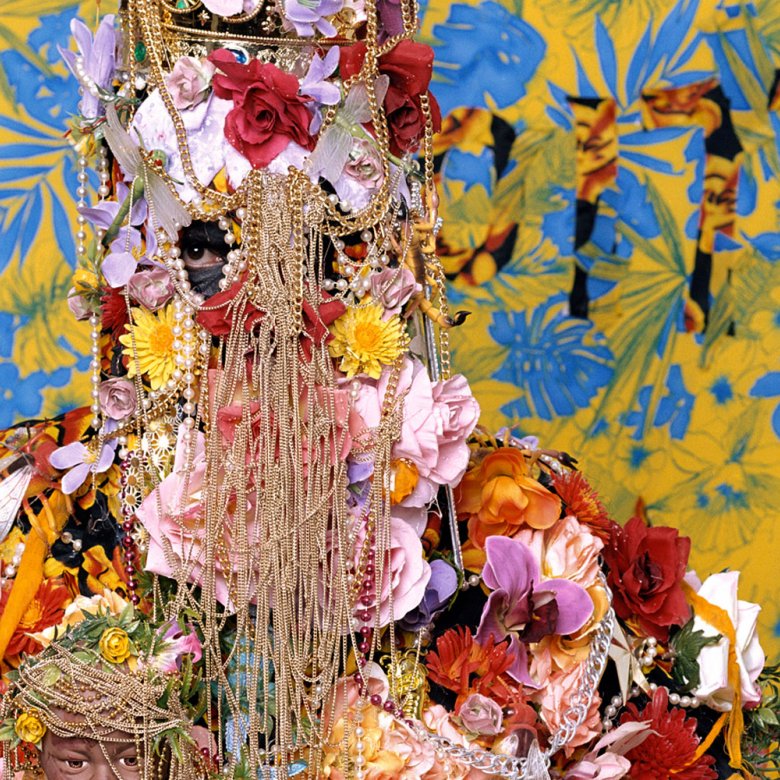 Figure covered in flowers and gold chains with just eyes showing through.