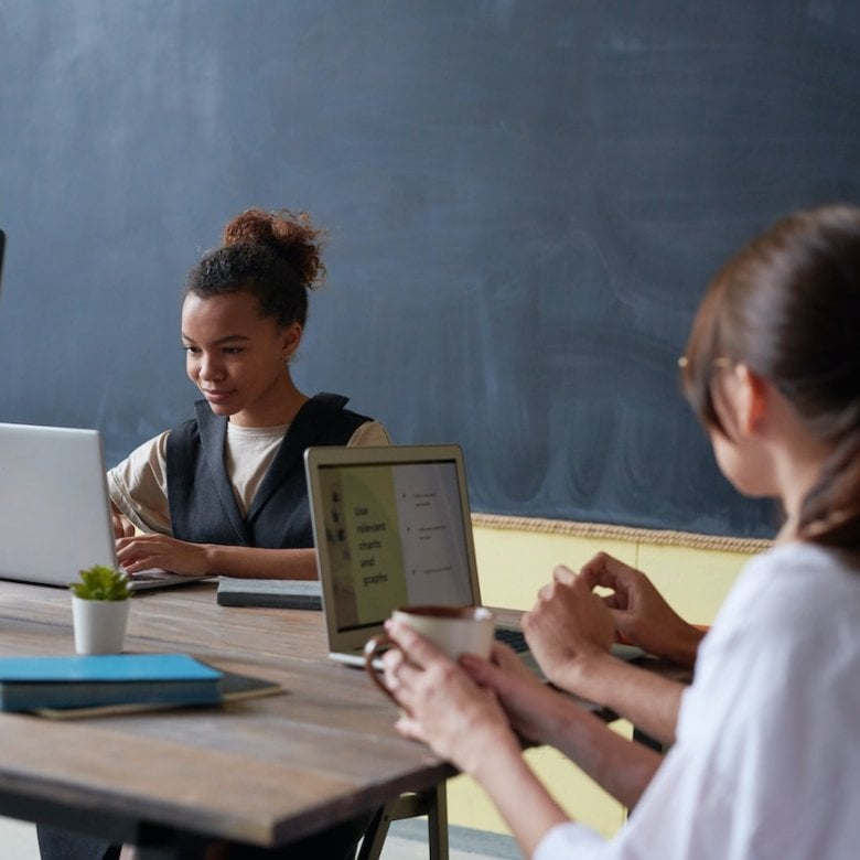 Three people studying at laptops on a table with a notebook and plant