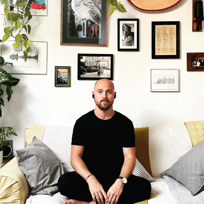 Graduate Thomas Young sitting cross-legged on a sofa with a wall of artwork and photos behind him