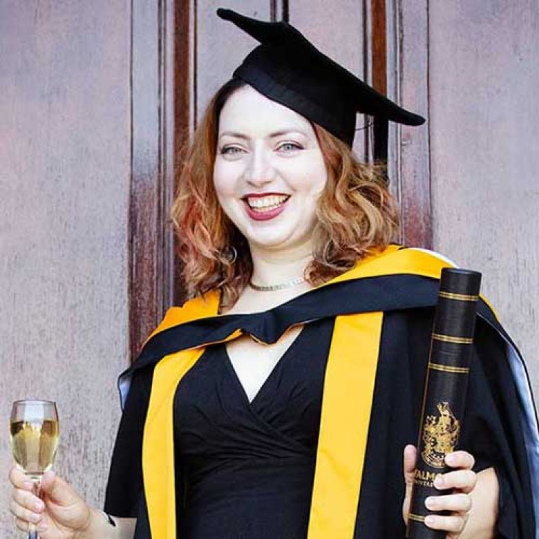 A graduate wearing her cap and gown, holding her degree certificate and a glass of wine