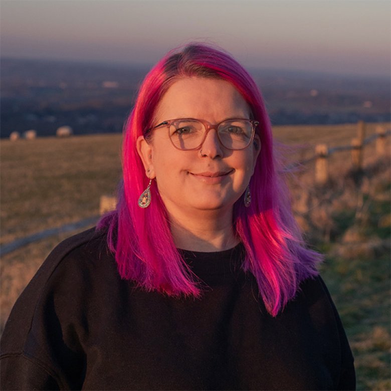 A headshot of a woman with pink hair and glasses