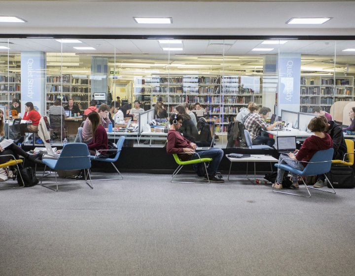 Students sat on low chairs working in front of large window looking through to the library on Penryn campus.