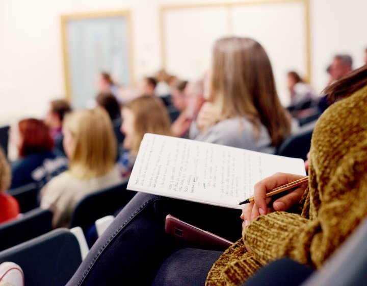 A person sitting in a lecture theatre with a notebook and pen in hand