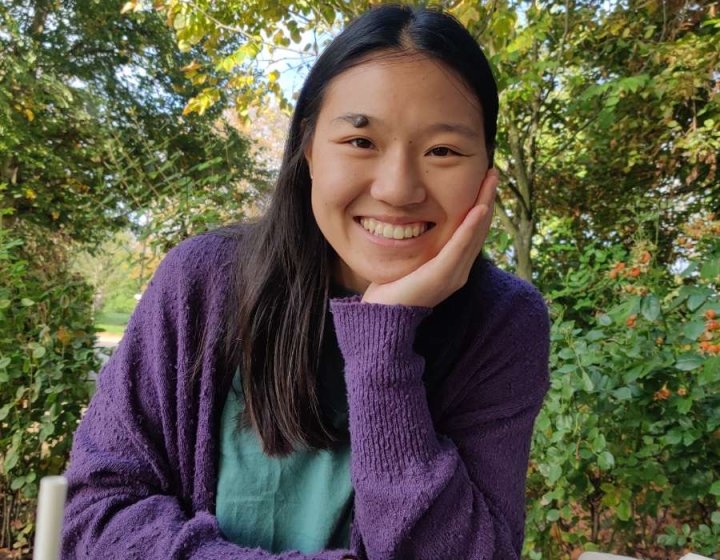 Female student wearing purple jumper 4smiling while sitting at wooden bench with leafy trees in the background.