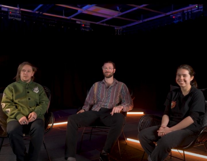 Photo of three people sitting on chair looking towards the camera. On the left, an anglepoise lamp sits on top of a side table. In the background there are neon strip lights.