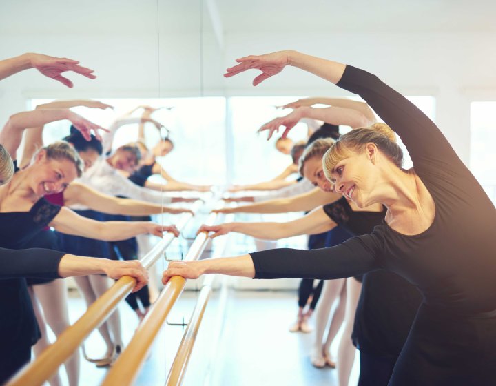A line of people dressed in black clothing doing ballet classes with a wall mounted barre looking at a mirror