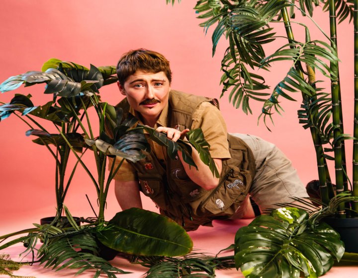 A person sat on the floor in jungle explorer clothing with plants around them against a light pink background