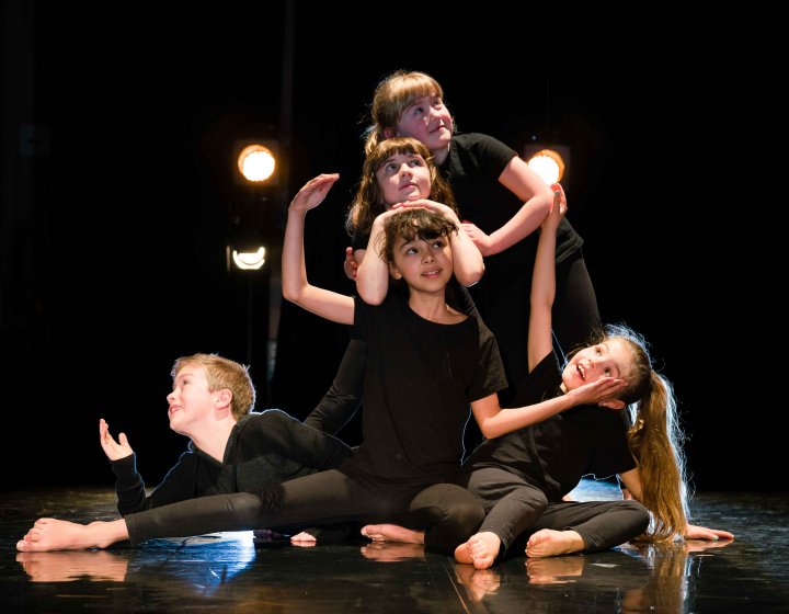 A group of mixed age young dancers posing on stage looking at the camera with some theatrical lighting in the background