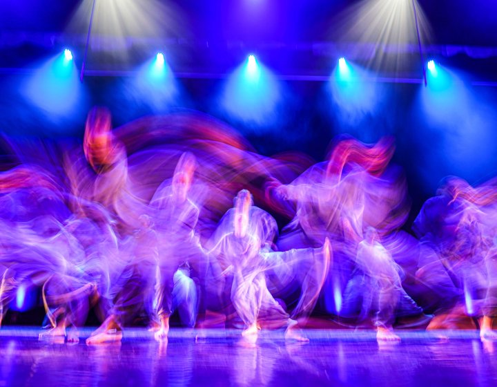 A blurred images of young dancers moving quickly across a large stage with blue lighting from above and purple lighting from the sides