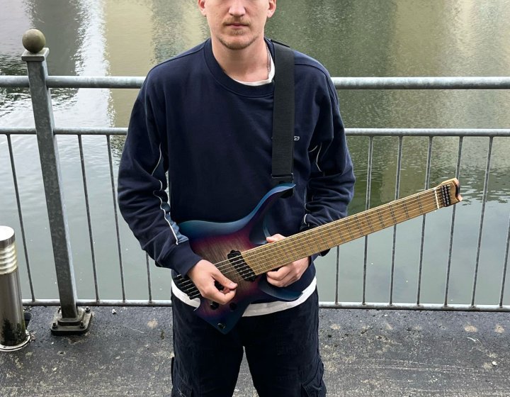 A solo performer's outside in front of a river holding a guitar