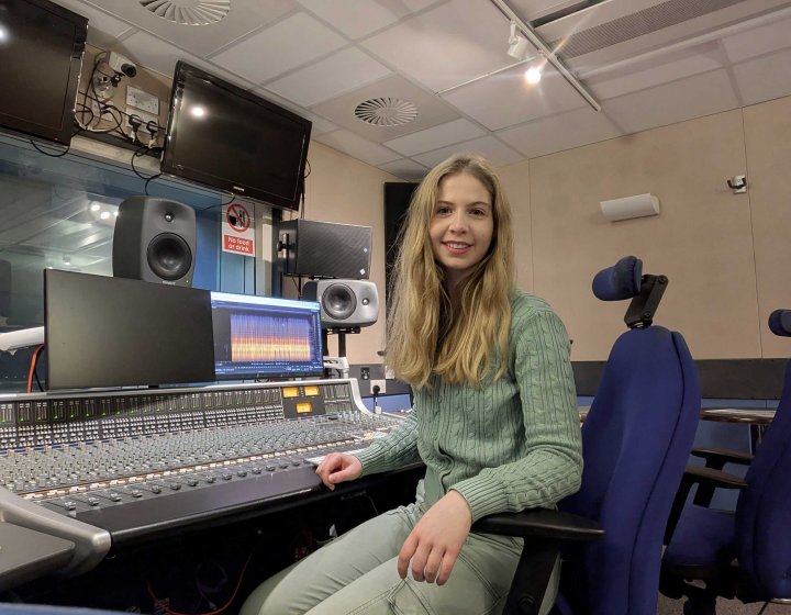 An image of Estella Hurlock sat on a blue chair wearing a green outfit in front of a large audio mixing desk