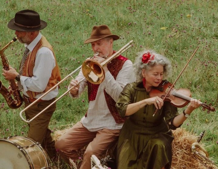 3 musicians playing a saxophone, trombone and violin in a field sat on a hay bale with grass in the background