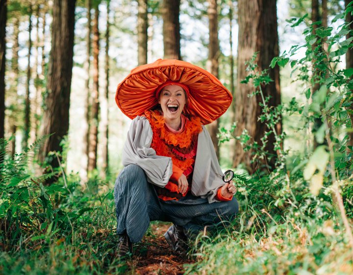 A person in the woods with a large orange fabric mushroom hat on