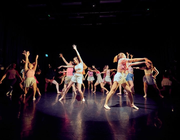 A large dance group ensemble performing on stage in a mixture of pink and stripy colour costumes