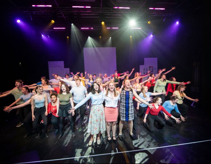 A large mixed performance ensemble on stage performing a finale group number under theatrical stage lighting