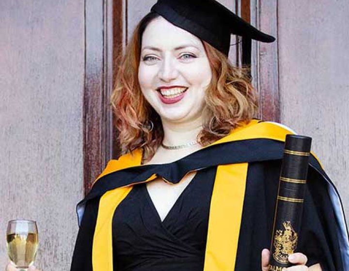 A graduate wearing her cap and gown, holding her degree certificate and a glass of wine