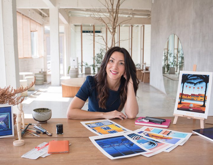 A head shot of illustrator Molly Maine, with long brown hair sitting at a wooden desk