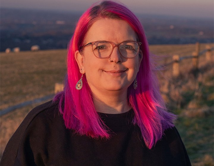 A headshot of a woman with pink hair and glasses