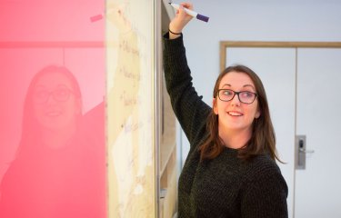 A woman wearing a black jumper, writing on a whiteboard with a pink panel