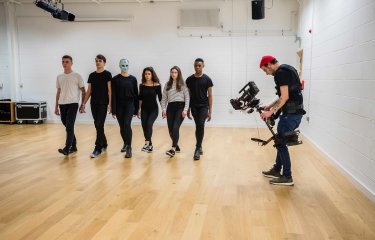 6 people walking in a line in a studio filmed by a lone cameraman 