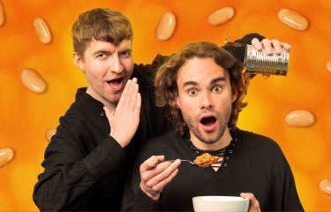 An image with two male performers, one whispering in the others ear whilst they are eating baked beans
