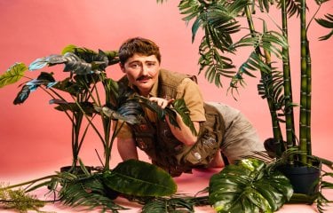 A person sat on the floor in jungle explorer clothing with plants around them against a light pink background