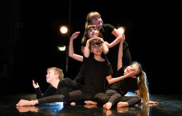 A group of mixed age young dancers posing on stage looking at the camera with some theatrical lighting in the background