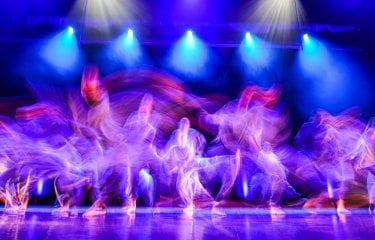 A blurred images of young dancers moving quickly across a large stage with blue lighting from above and purple lighting from the sides