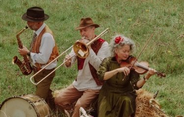 3 musicians playing a saxophone, trombone and violin in a field sat on a hay bale with grass in the background