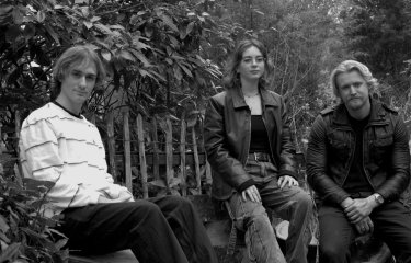 A black and white image of three musicians sat on a bench in a line.