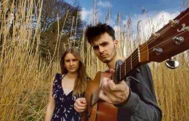 Two performers outside against a long grass background. The performer in the front of the image is holding a guitar