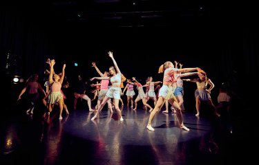 A large dance group ensemble performing on stage in a mixture of pink and stripy colour costumes