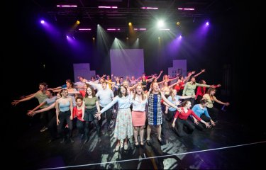 A large mixed performance ensemble on stage performing a finale group number under theatrical stage lighting