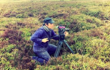 A filmmaker crouches behind his camera amongst shrubbery 