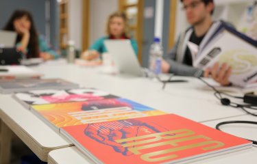 Design journals on a desk in the foreground, students working out of focus in the background.