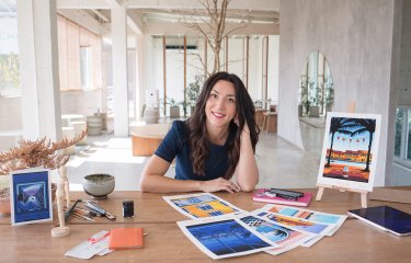 A head shot of illustrator Molly Maine, with long brown hair sitting at a wooden desk