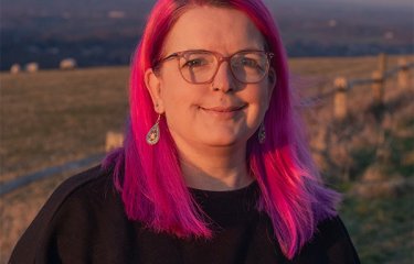 A headshot of a woman with pink hair and glasses