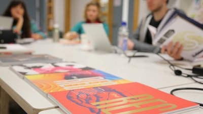 Design journals on a desk in the foreground, students working out of focus in the background.