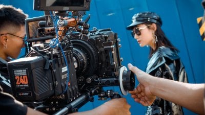 A man controlling a RED camera filming a women wearing sunglasses, a leather hat and jacket