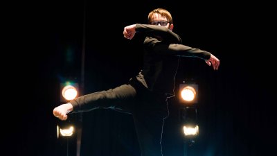 A young person making a dance pose towards the camera with stage lights shining in the background