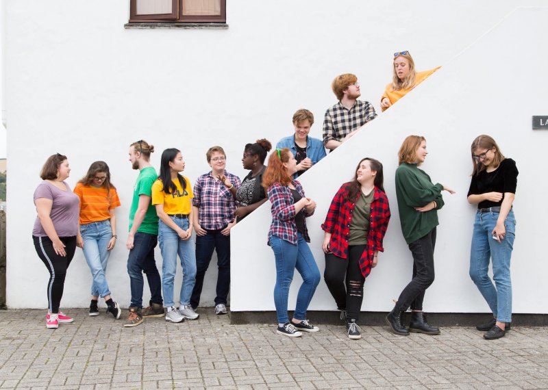 13 Falmouth University students in different coloured tops stood against a white wall and on a white staircase.