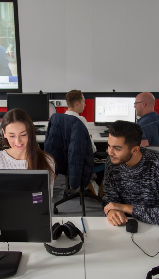 Students working together in newsroom, news playing on screens behind them.