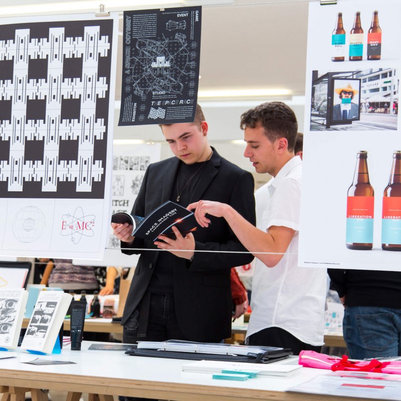 Students looking through a book, design work hanging from a line in front of them.