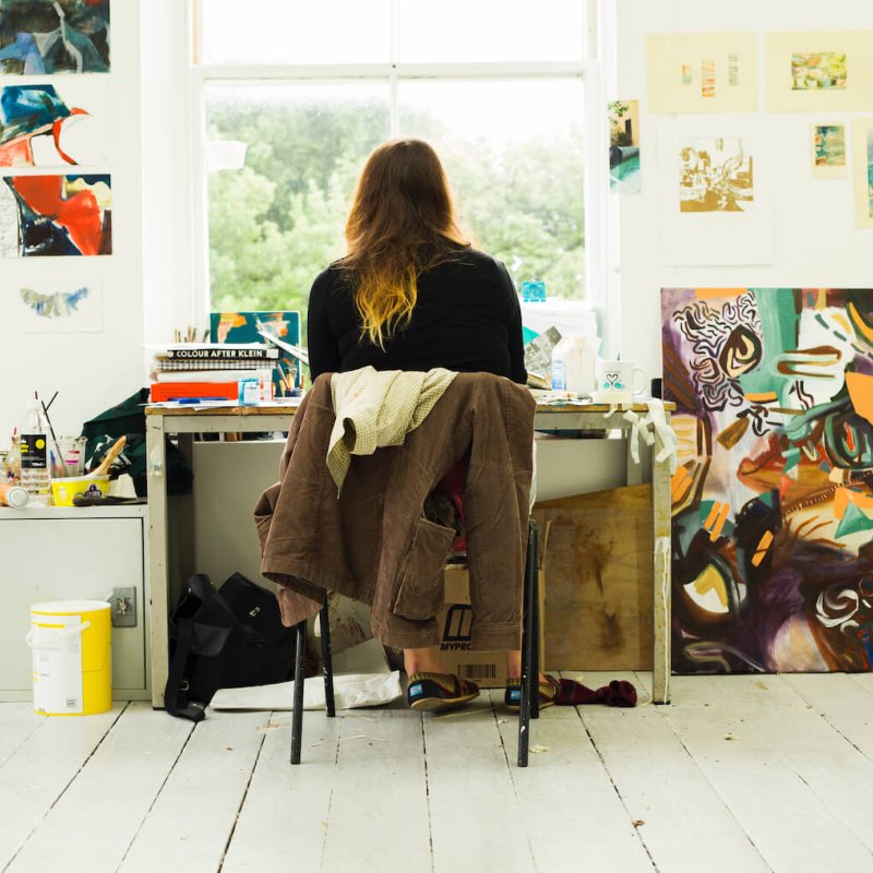 Back of student sitting at desk in front of window surrounded by artwork and paintings.