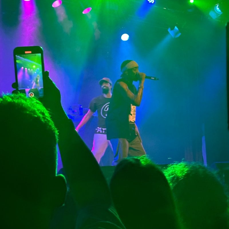 Two men performing on a stage with blue lights and person filming on smartphone
