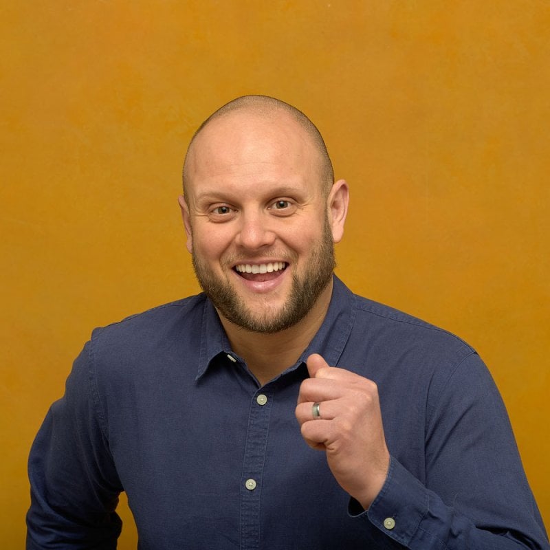 Headshot image of comedian Tom Glover. Tom has a bald head, is smiling at the camera and wearing a blue shirt against a mustard colour backdrop